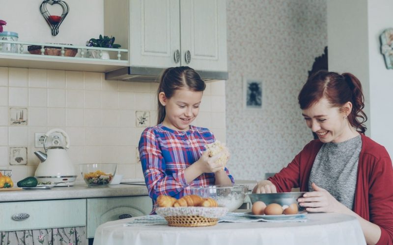 Little cute girl helping her mother in the kitchen stirring dough for cookies into bowl. Mom have fun smearing daughter nose with flour and they laughing . Family, food, home and people concept