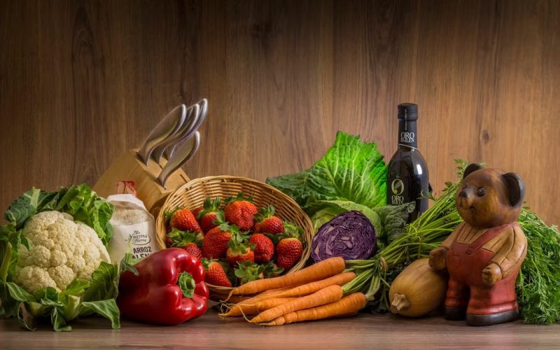 Arrangement of fresh vegetables and fruits on a wooden table with kitchen essentials.